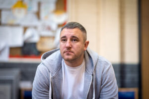 A middle-aged white man with grey hair sits down wearing a grey hoodie