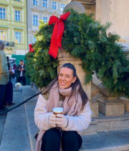 FullSizeRender A person sat outside on a step in front of a festive wreath wearing a coat and scarf, holding a drink.