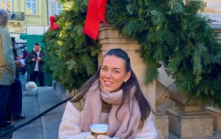 A person sat outside on a step in front of a festive wreath wearing a coat and scarf, holding a drink.