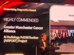 A big screen saying Modernising Diagnostics Award Highly Commended Greater Manchester Cancer Alliance AI Radiology PriORiTisation (AIRPORT) Project ands man on stage announcing the accolade