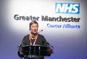 Jo Taylor web story tribute The late Jo Taylor a white woman with short hair standing on a stage at a lectern giving a talk with the words NHS Greater Manchester Cancer Alliance behind her