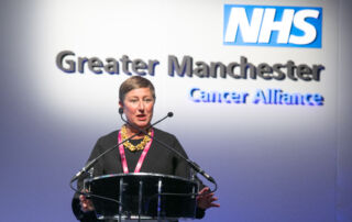 The late Jo Taylor a white woman with short hair standing on a stage at a lectern giving a talk with the words NHS Greater Manchester Cancer Alliance behind her
