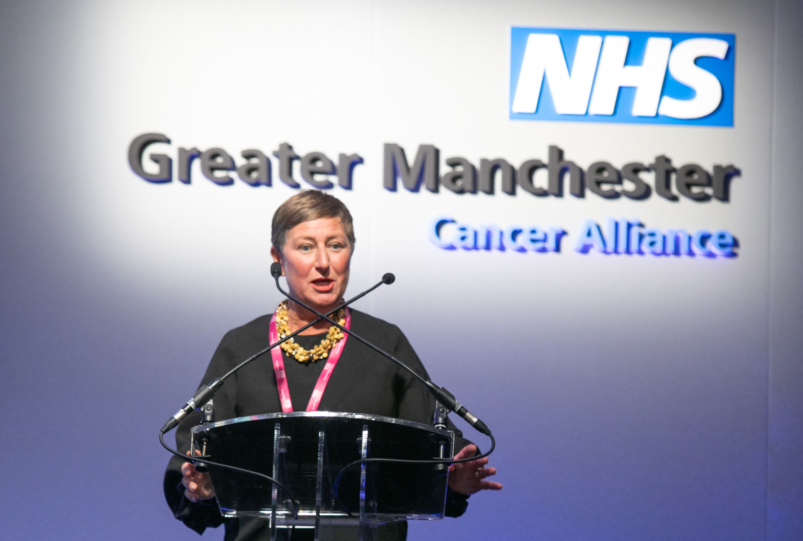The late Jo Taylor a white woman with short hair standing on a stage at a lectern giving a talk with the words NHS Greater Manchester Cancer Alliance behind her