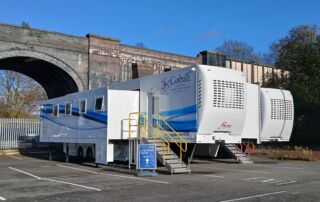 Two white trucks housing a mobile lung cancer screening service in front of an arch in a car park
