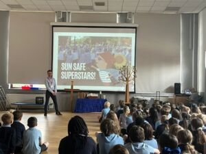 An hall at a school full of children during an assembly. A teacher is stood at the front of the hall in front of a project screen showing a presentation on sun safe superstars.