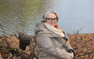 Gillian Glynn, a white woman in her 60s, sits on a park bench by a lake