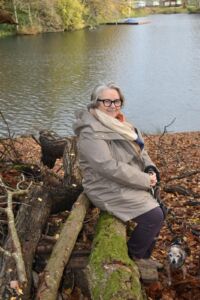 Gillian Glynn, a white woman in her 60s, sits on a park bench by a lake with Autumn leaves at her feet