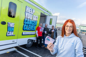 Four people stand in front of a brightly coloured green roadshow unit with the words: "Help You Get Cancer Clever" on the side. A woman with red hair stands towards the front holding a leaflet with the words Get Cancer Clever on the front. Three men stand a little further behind near an open door on the roadshow