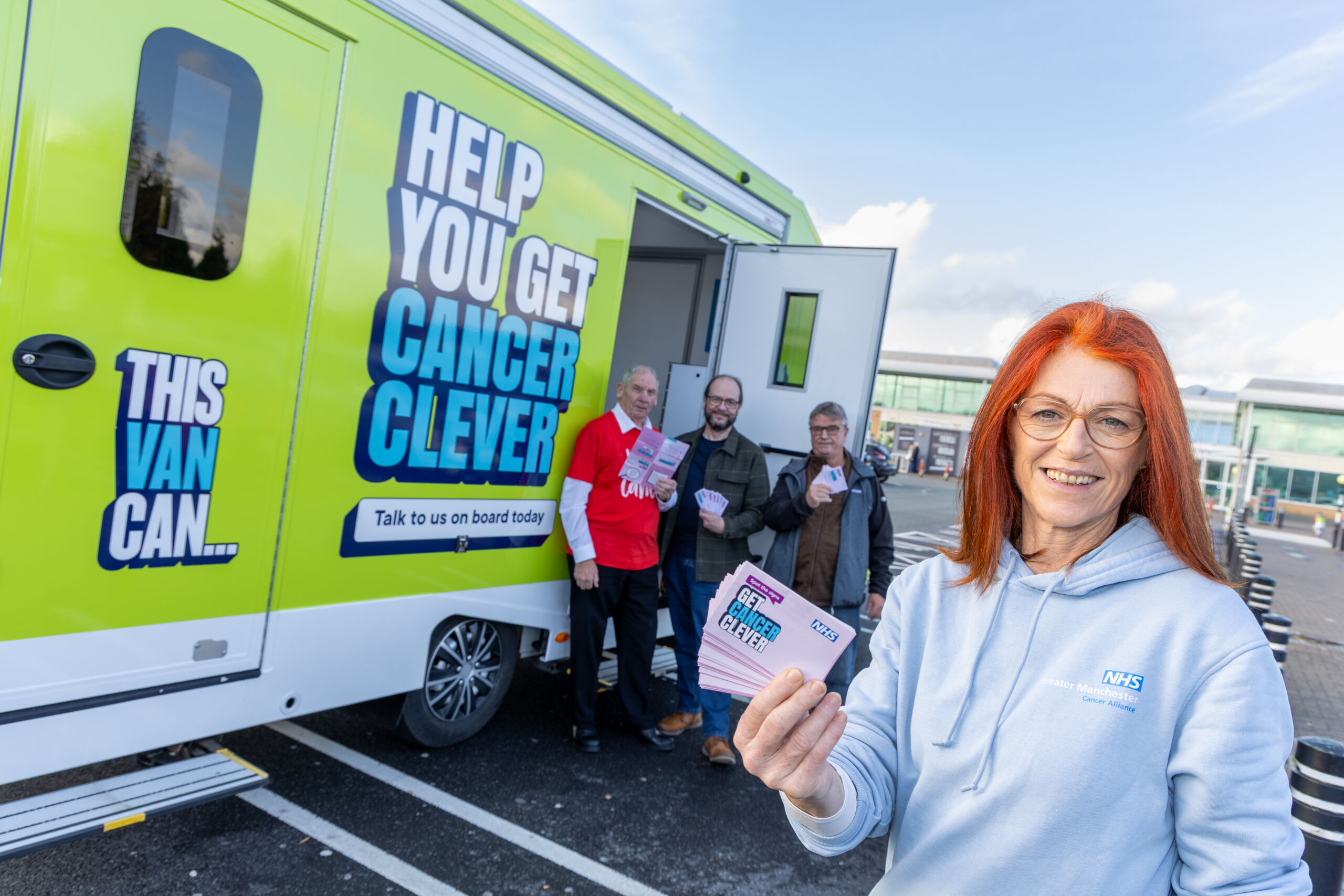 Four people stand in front of a brightly coloured green roadshow unit with the words: "Help You Get Cancer Clever" on the side. A woman with red hair stands towards the front holding a leaflet with the words Get Cancer Clever on the front. Three men stand a little further behind near an open door on the roadshow