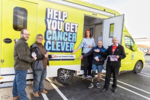 Four people stand in front of a brightly coloured green roadshow unit with the words: "Help You Get Cancer Clever" on the side. 