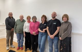 A group of seven men and woman who are patient carers standing against a white wall in casual clothes with two members of staff from Made by Mortals