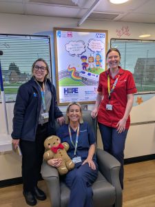Three people in healthcare uniforms are smiling in a clinical room on board the mobile cancer care unit. One person is sitting in a chair holding a gingerbread plush toy, while the other two stand beside them. Behind them, there's a colourful NHS poster with cartoon characters.