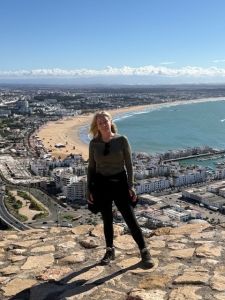 A full length image of a woman Sharon Marriot with blonde hair wearing a black top and trousers standing on hill top above a beach