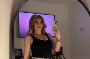 A young woman with blonde hair wearing a black vest top takes a selfie of herself in front of a mirror