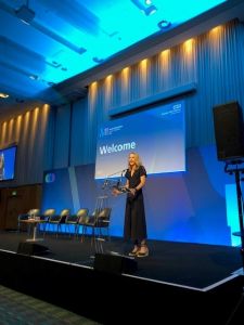 Olympian Paula Radcliffe talking on stage at a lectern with the words Welcome Greater Manchester Cancer Alliance in the background