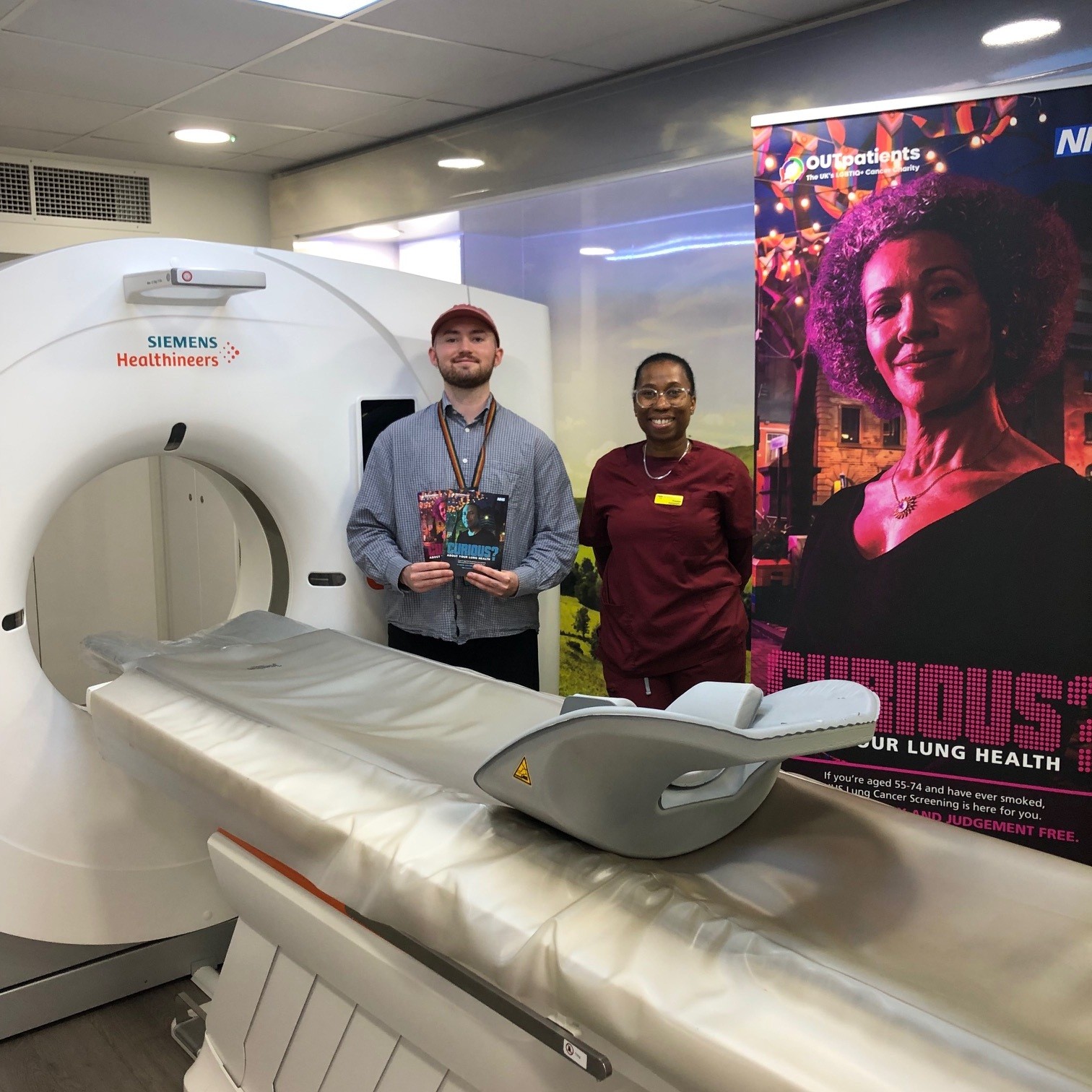 Ezra Johnson Communications and Public Affairs Manager from OUT Patients stands with a radiologist with a scanner inside the mobile NHS Lung Cancer Screening clinic a banner in the background aimed at the LBGTQI+ community asks people to Get Curious About Their Lung Health