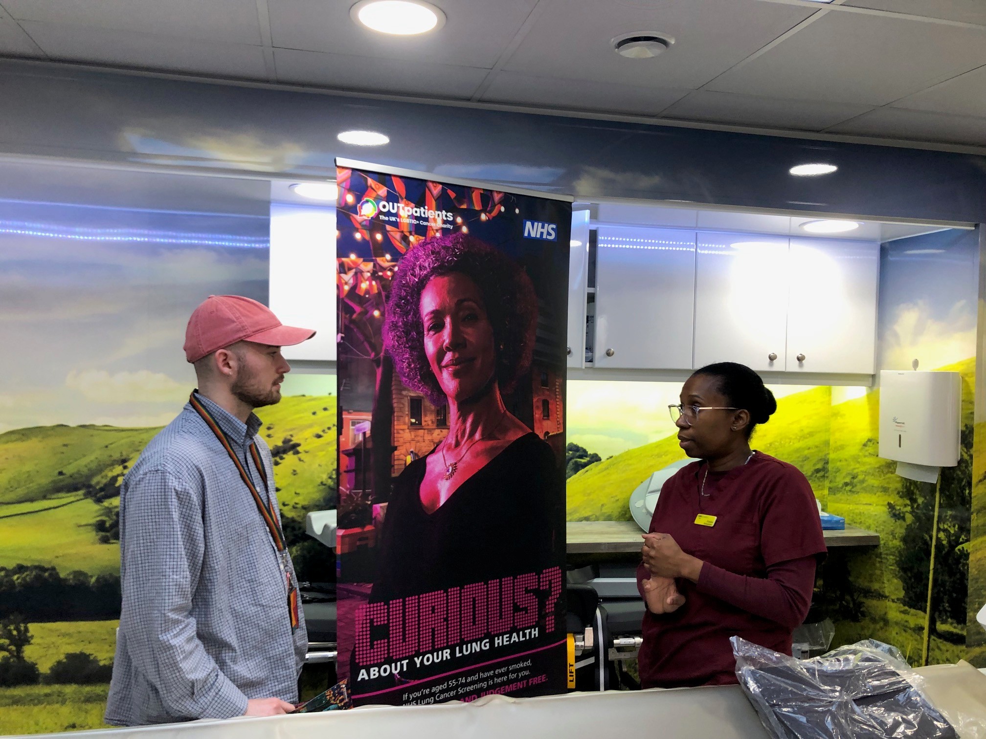 Ezra Johnson Communications and Public Affairs Manager from OUT Patients chats with a radiologist with a scanner inside the mobile NHS Lung Cancer Screening clinic a banner in the background aimed at the LBGTQI+ community asks people to Get Curious About Their Lung Health