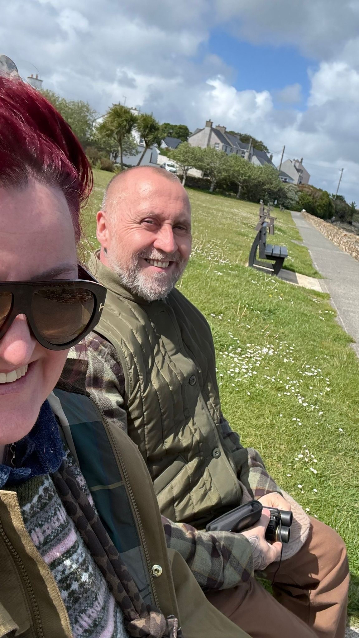 Gary a white man in his 50s with balding short hair sits on a park bench with his wife Nuala who has dark brown hair and wears sunglasses and is taking a selfie of them both