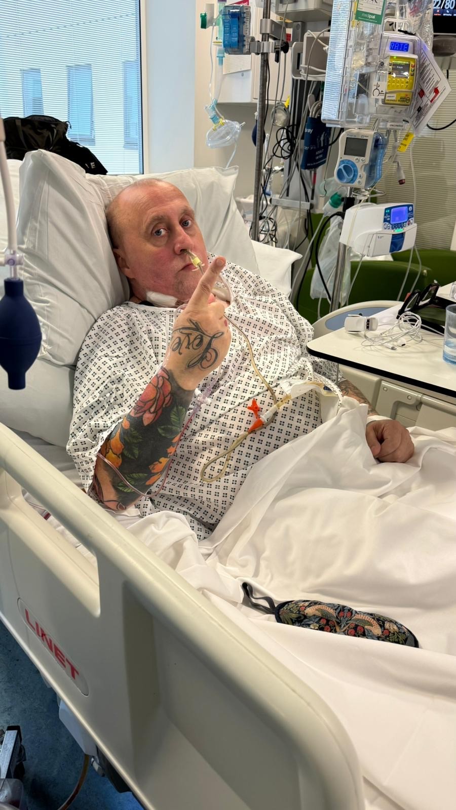 Gary a white man in his 50s with balding short hair lies sat up in a hospital bed wearing a hospital gown and pointing at the camera