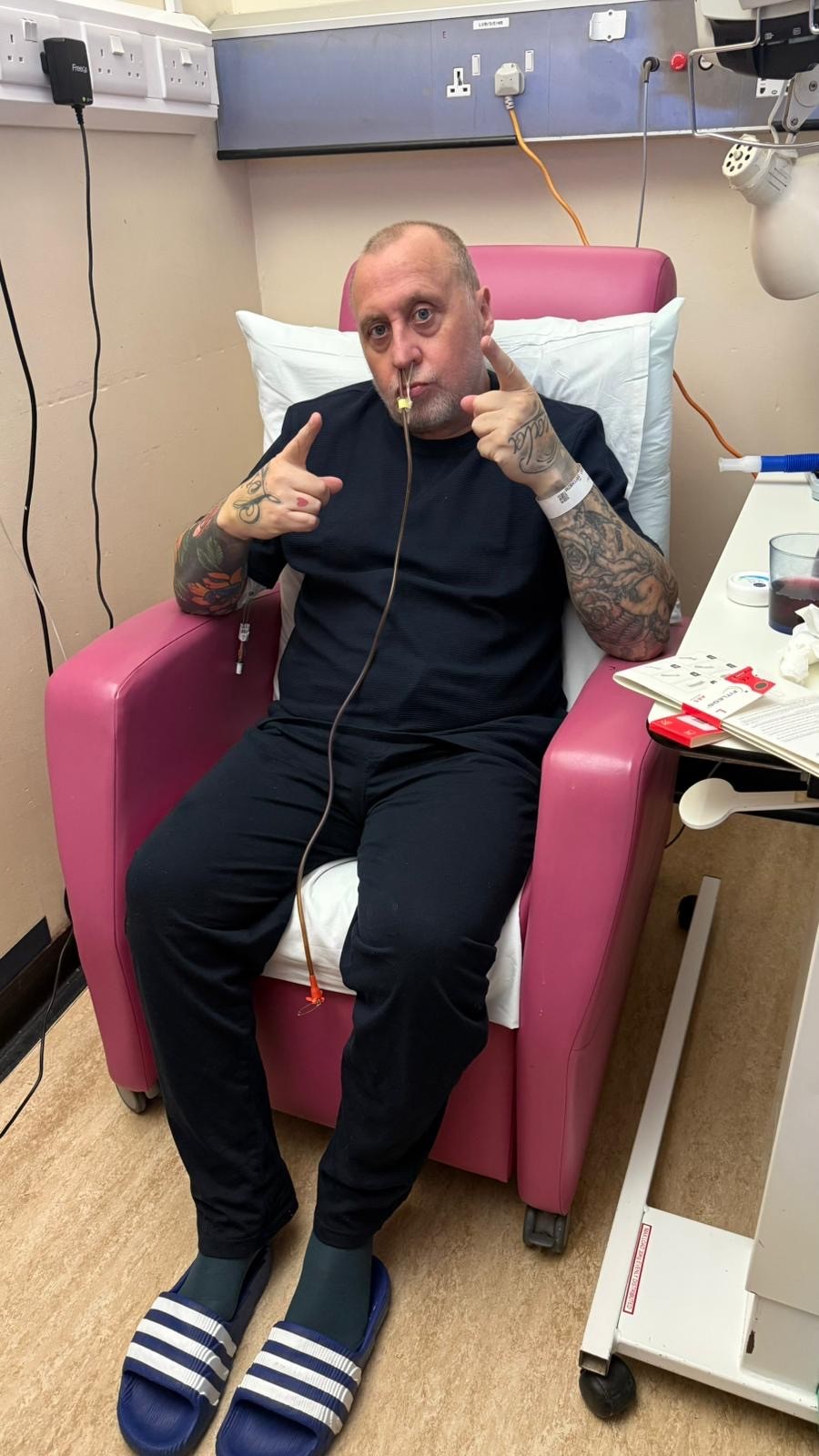 Gary a white man in his 50s with balding short hair sits in a hospital red chair next to hospital equipment while receiving his cancer treatment and makes pointing signs with his hands to the camera