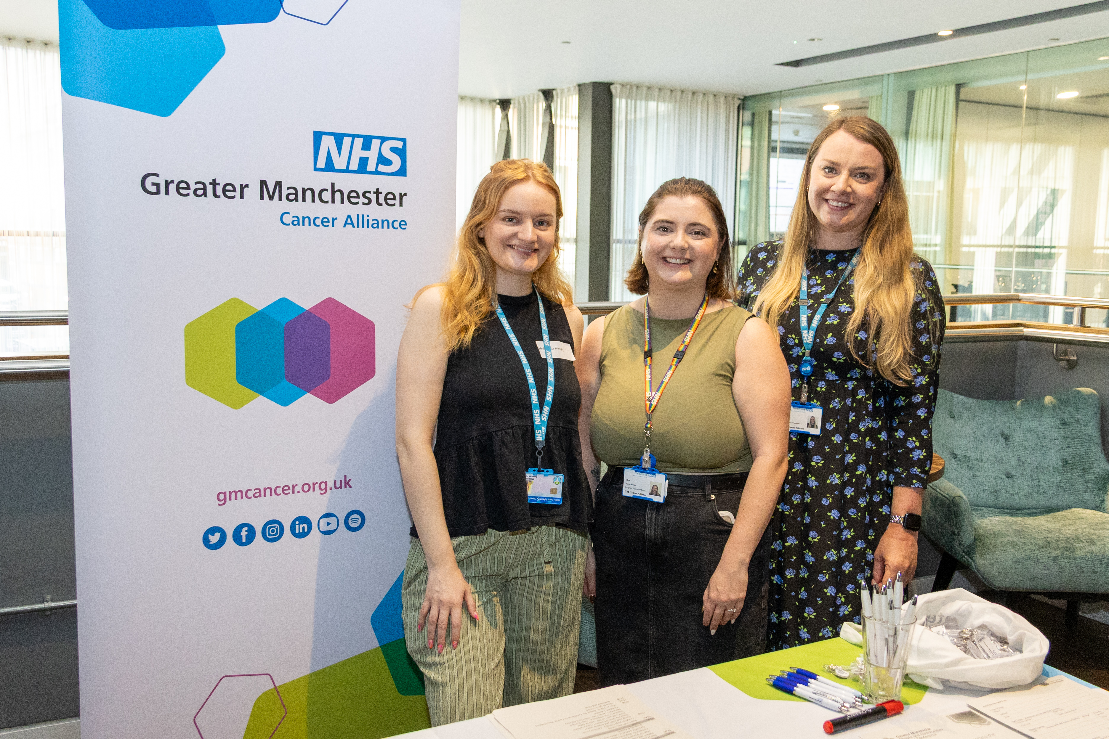1 Rebecca, Ellen and Pippa from the GM Cancer Alliance team stands smiling next to a banner for the Alliance. A table with papers and pens is in front of them. The setting appears to be a modern conference or event space.