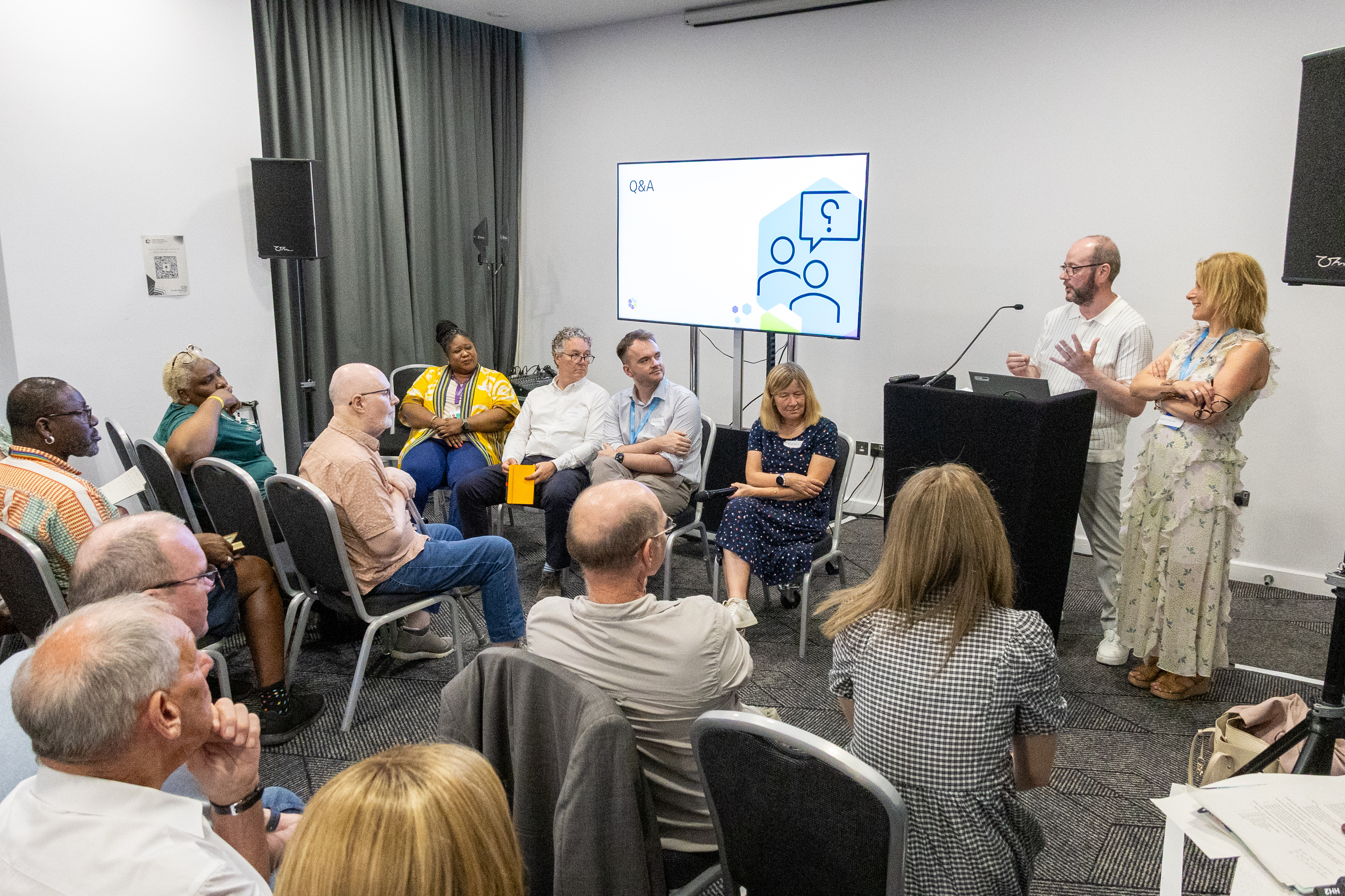 11 A group of people are seated in a semi-circle in a conference room, engaged in a discussion. Two individuals are standing at the front near a screen displaying "Q&A" with an icon. The room has neutral-coloured walls and curtains.