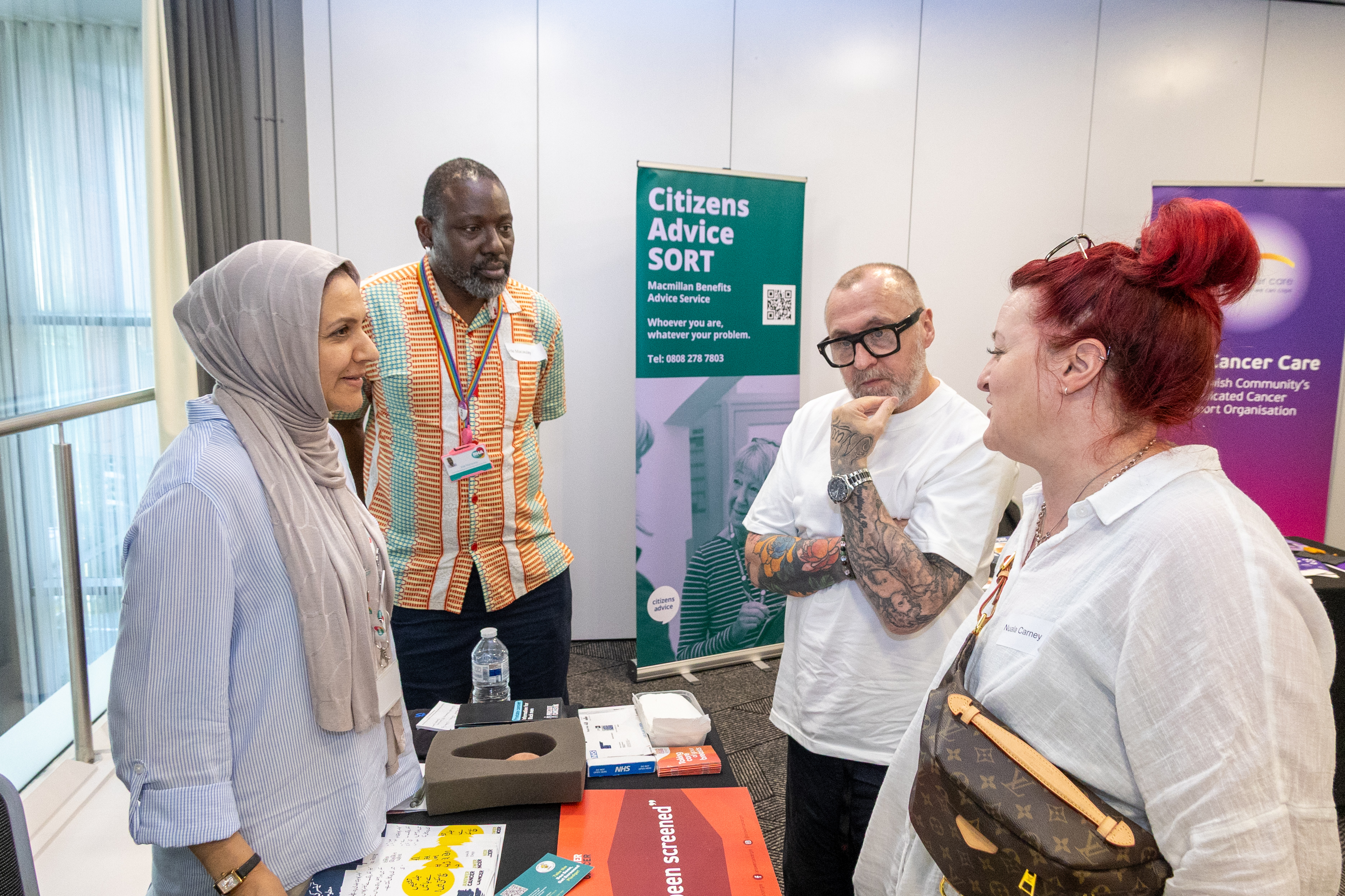 12 A group of people are having a conversation at an event. They are standing near a table with informational materials. In the background, there are banners for "Citizens Advice SORT" and "Cancer Care.