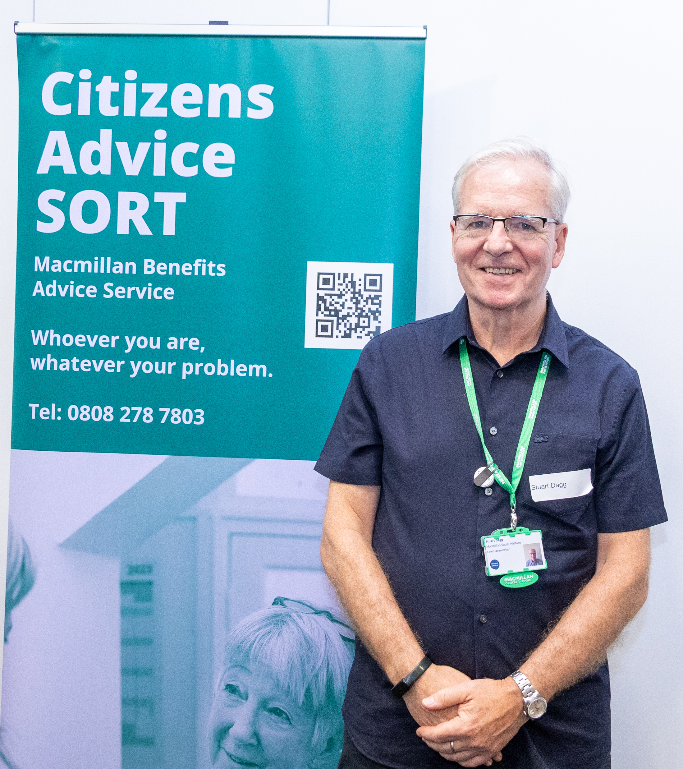 13 The image shows an individual standing beside a banner for the Citizens Advice SORT, Macmillan Benefits Advice Service. The banner includes contact information and a QR code. The person is wearing a name badge and an identification lanyard.