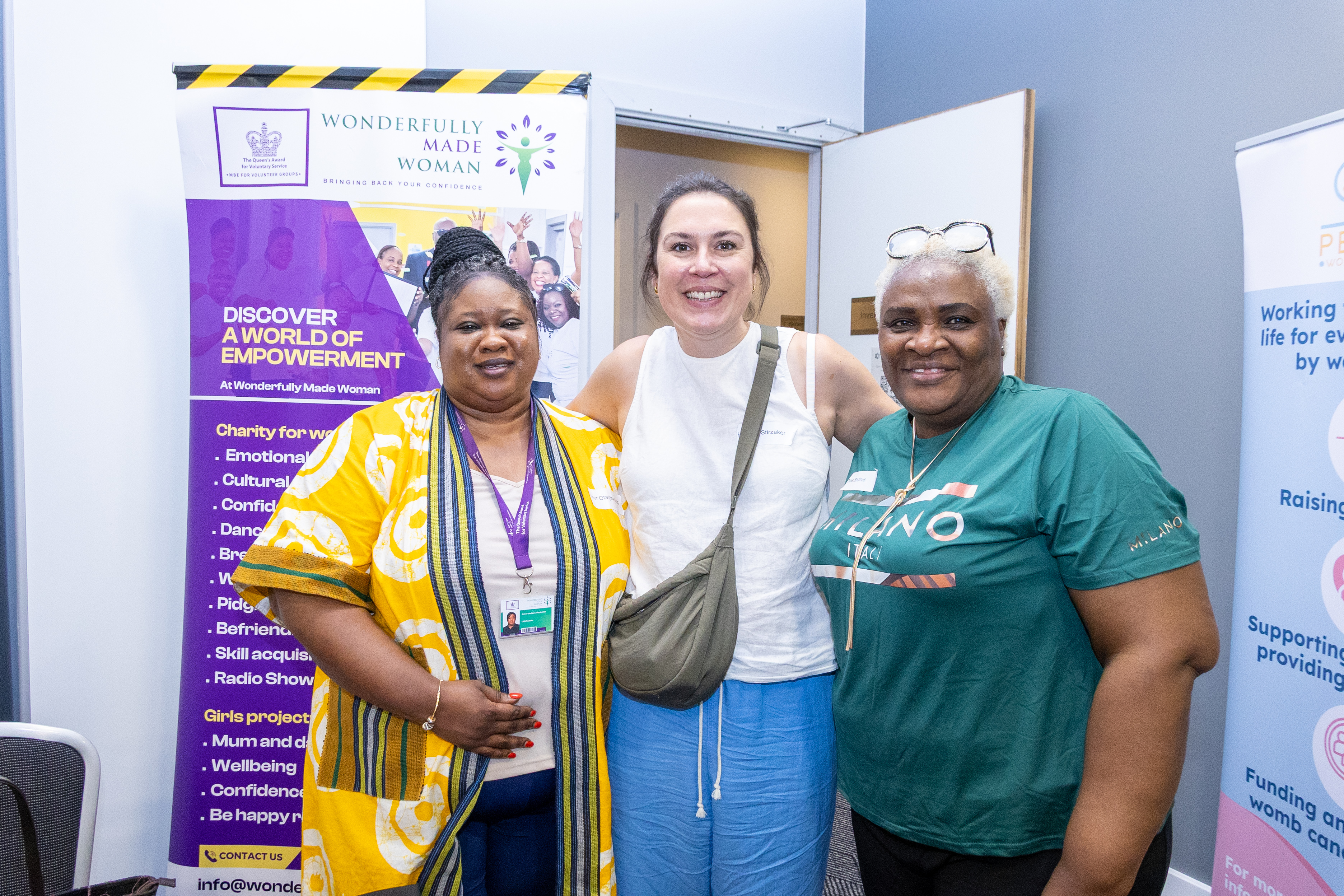 14 Three individuals are smiling in front of promotional banners for “Wonderfully Made Woman.” They are standing closely together in a room. The person on the left is wearing a yellow patterned outfit, the middle individual is dressed in a white top and blue pants, and the person on the right has a green shirt. The background features other branded materials and a doorway.