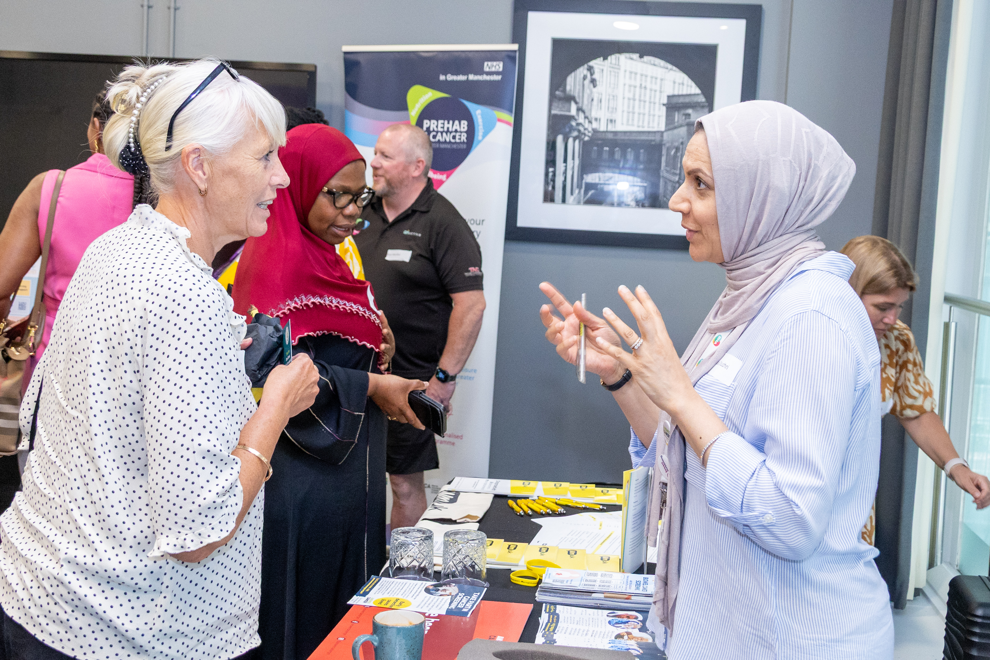 15 A group of people are gathered around an informational table at an event. There are brochures and materials related to cancer awareness on the table. The setting appears to be indoors, and there is a framed black and white photo of a building on the wall in the background. One person is speaking animatedly, gesturing with their hands.