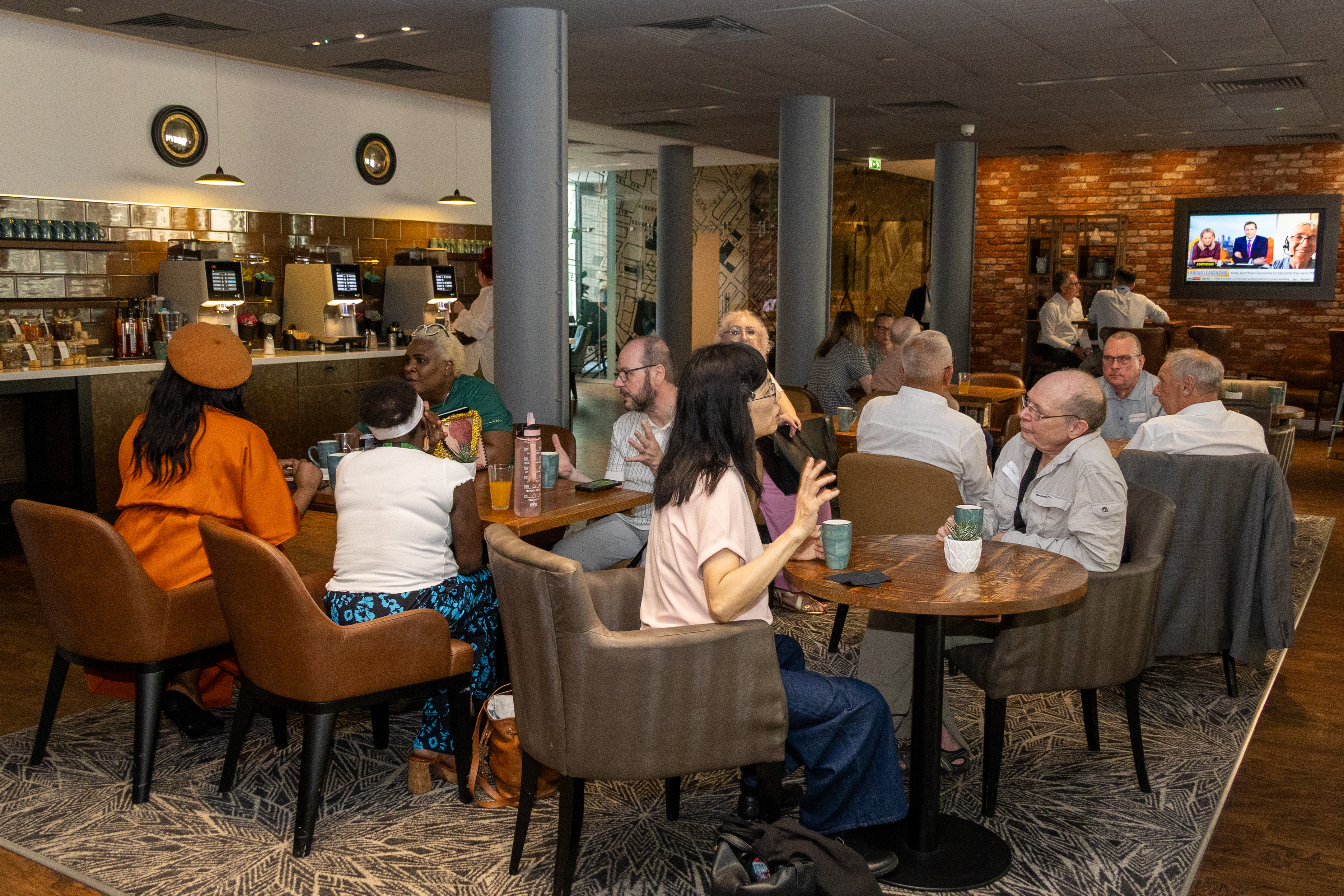 2 A group of people are seated around tables in a cosy conference break area with a mix of brown chairs and sofas. Several people are engaged in conversation, while others are seated alone. A flat-screen TV on the brick wall displays a news program, and the counter at the back has coffee machines and pastries. The atmosphere appears lively and social.