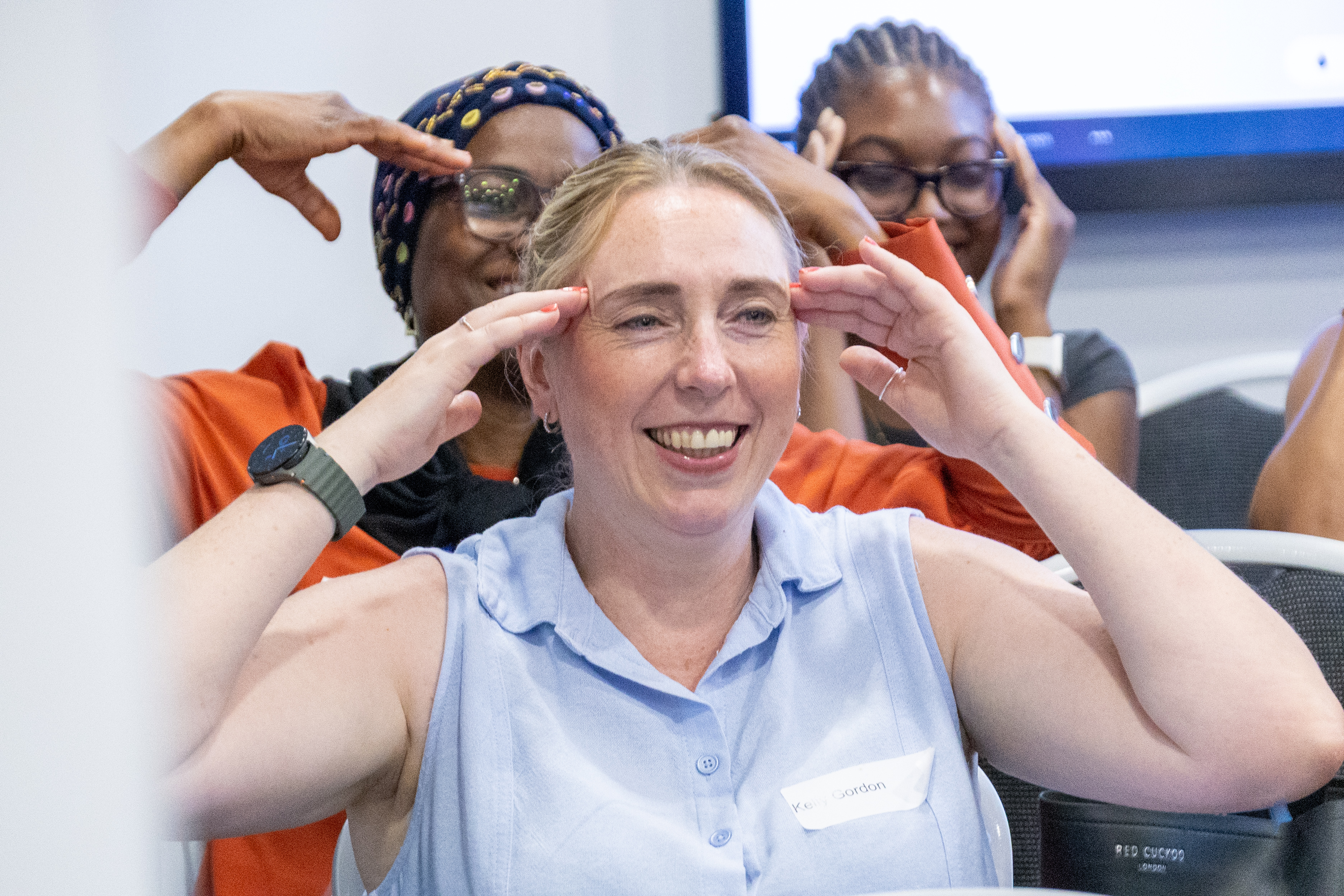 22 A group of attendees taking part in a guided chair yoga session all smiling with hands on their head. The image is focused on a woman in a blue top wearing a health watch
