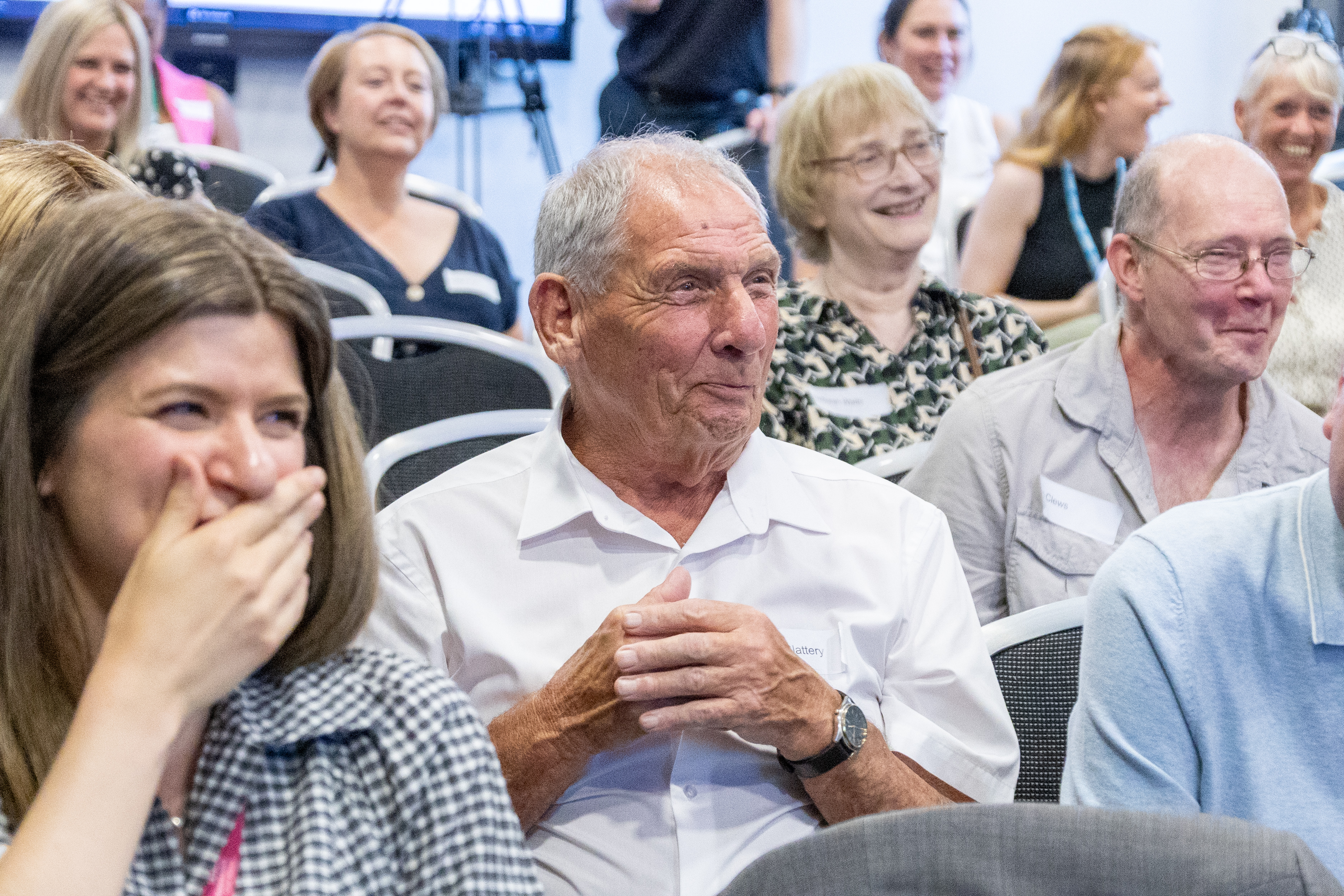 23 A group of attendees laughing and smiling engaged in a session at a conference with focus on a male in a white shirt
