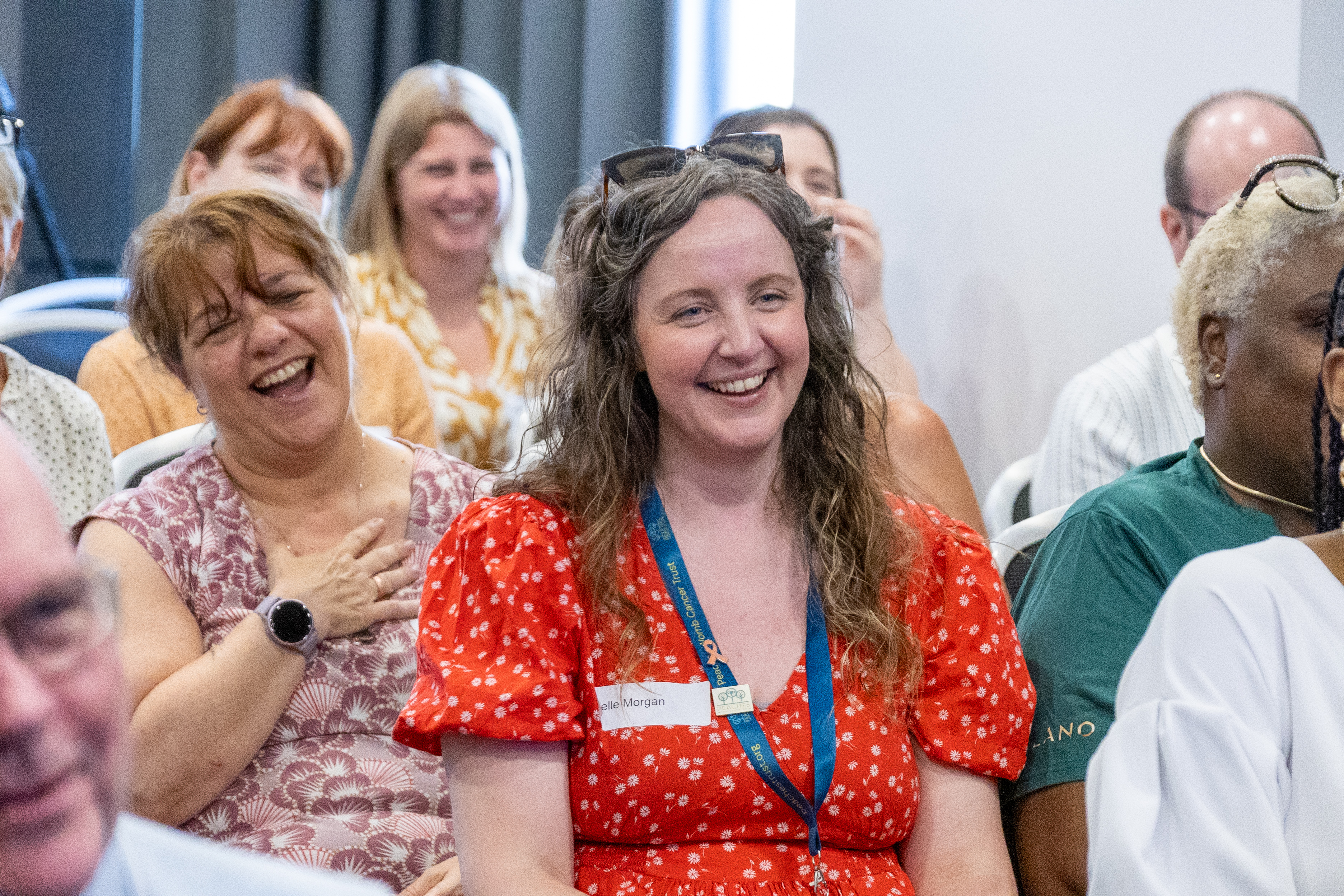 24 Delegates sat at a conference laughing with the focus on Joelle Morgan from Peaches Womb Cancer Charity wearing a floral red dress smiling in her seat