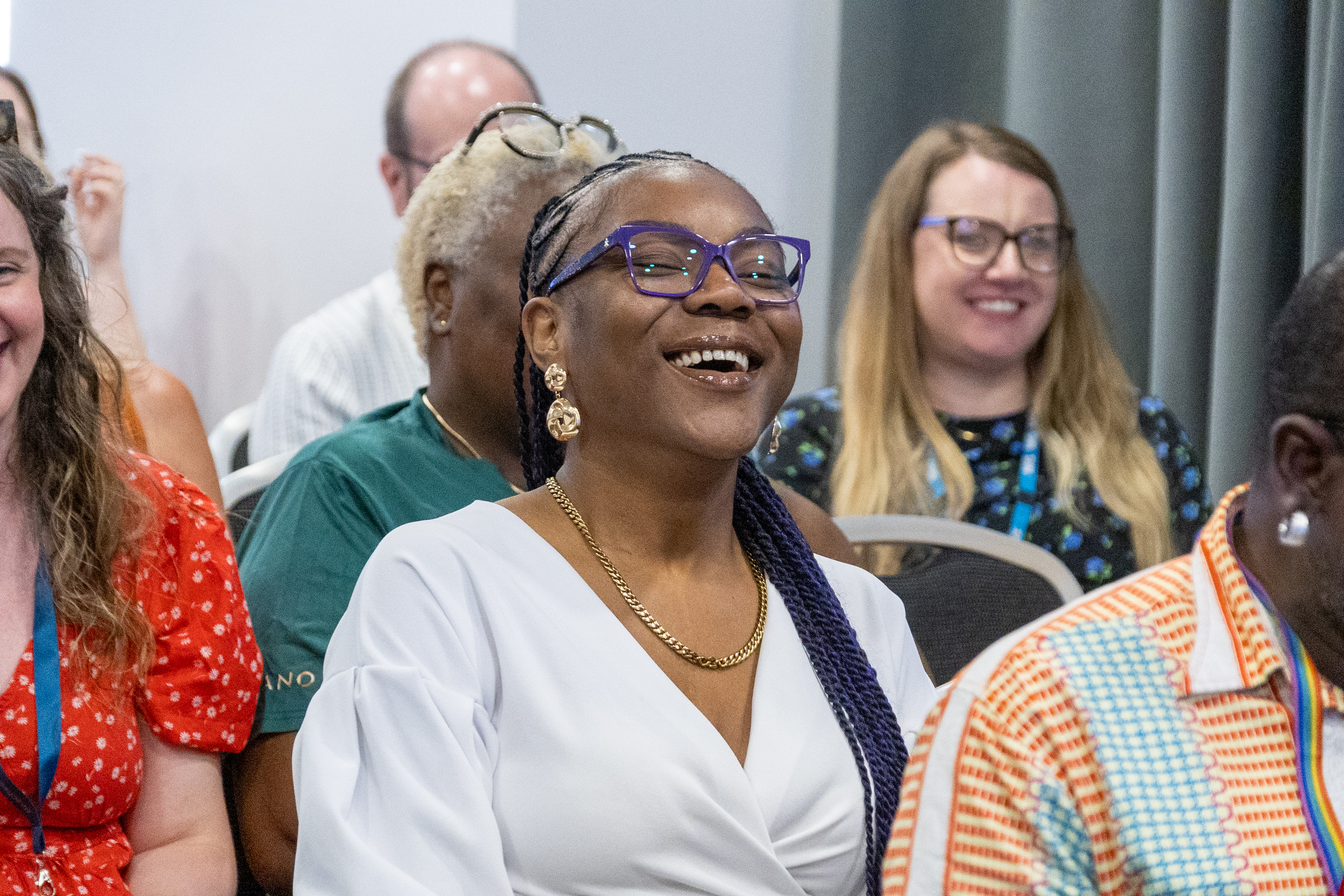 25 Close up of Maria a patient representative sat in the audience of a conference smiling wearing a white top and purple glasses