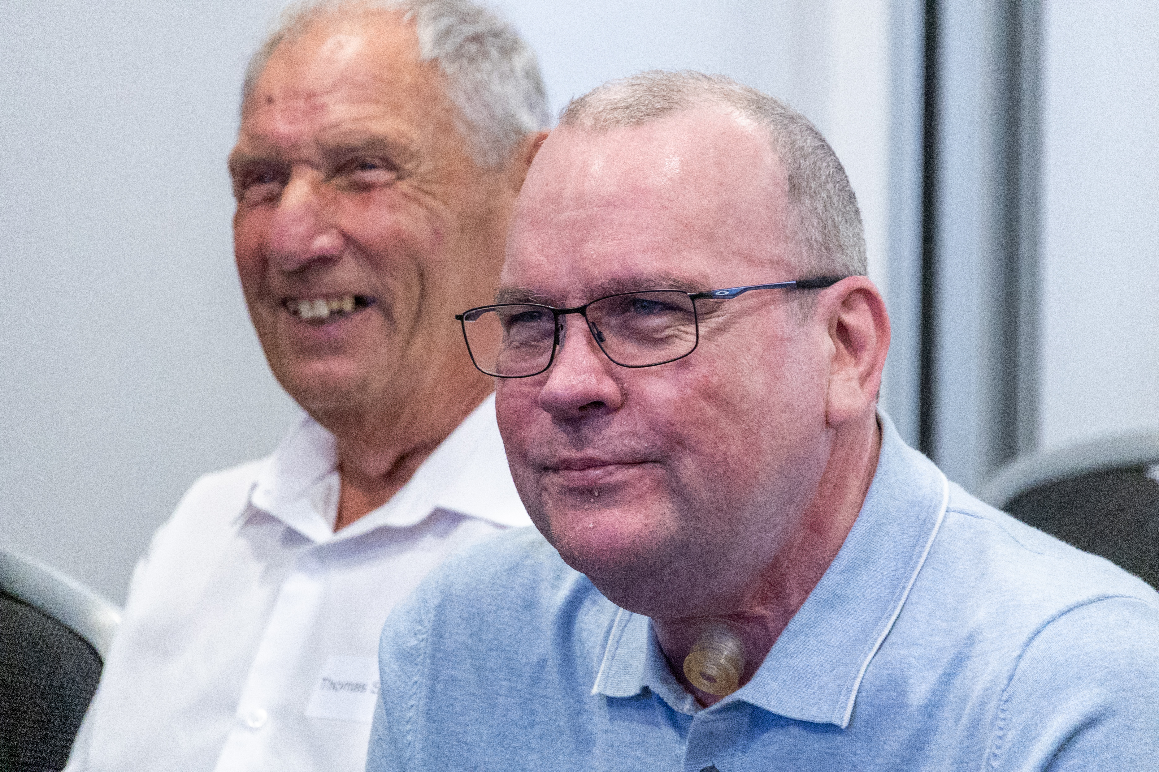30 Two people are sitting and smiling. The person in the foreground is wearing glasses and a light blue shirt, and has a medical device on their neck