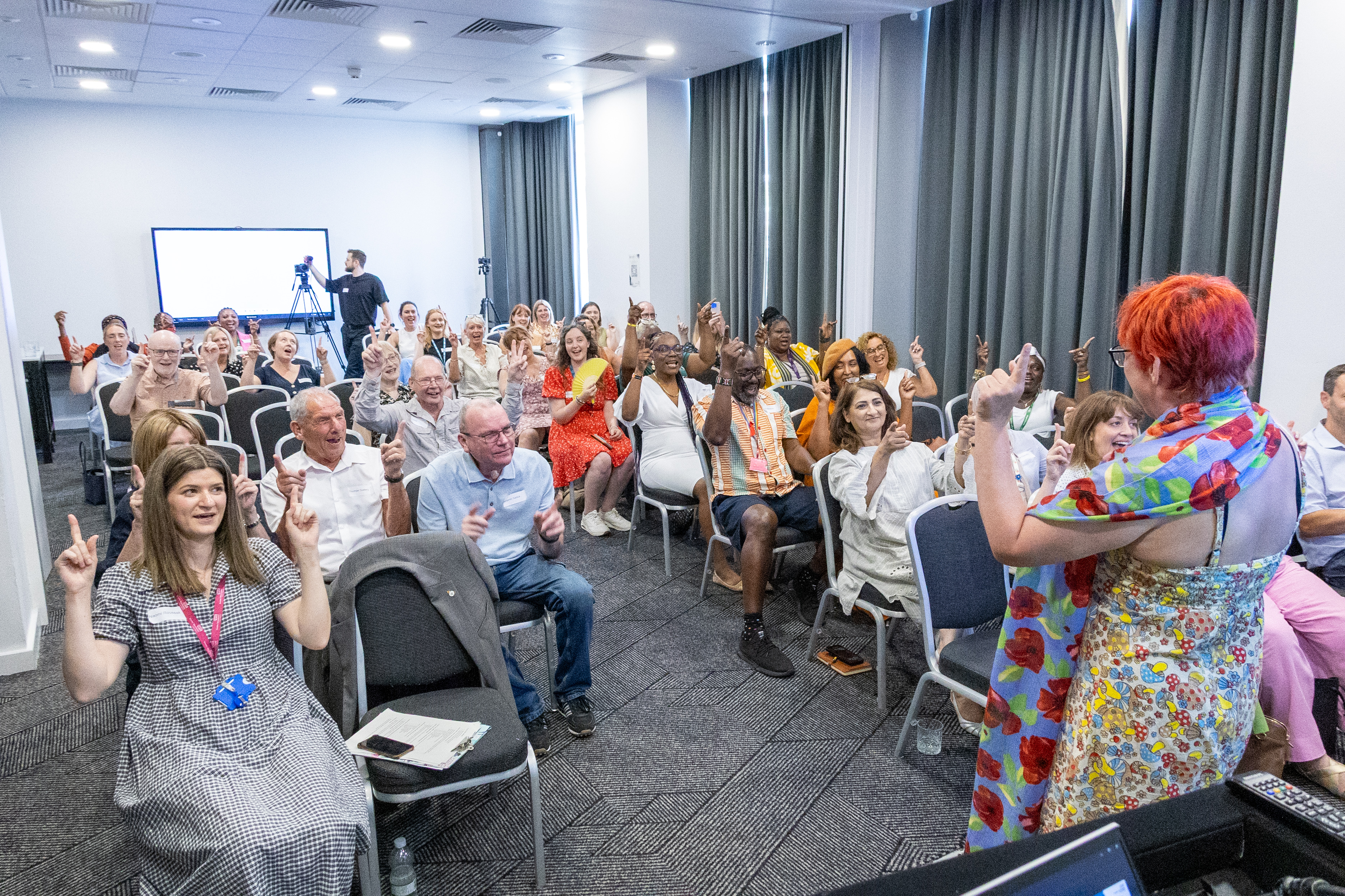 32 Delegates at the conference seated and joining in on a laughter yoga therapy session with arms up in the air and enthusiastic expressions on faces