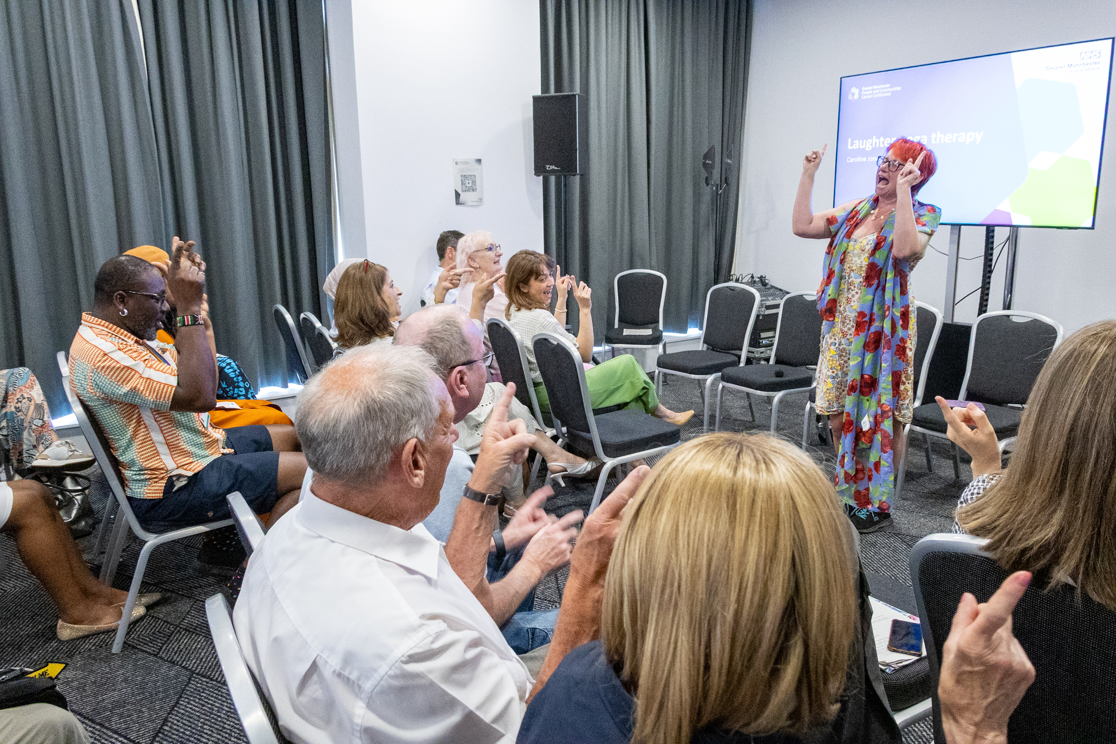 34 Caroline Jone stood at the front with arms in the air as she facilitates a laughter yoga session with the backs of delegates heads all sat joining in on the session