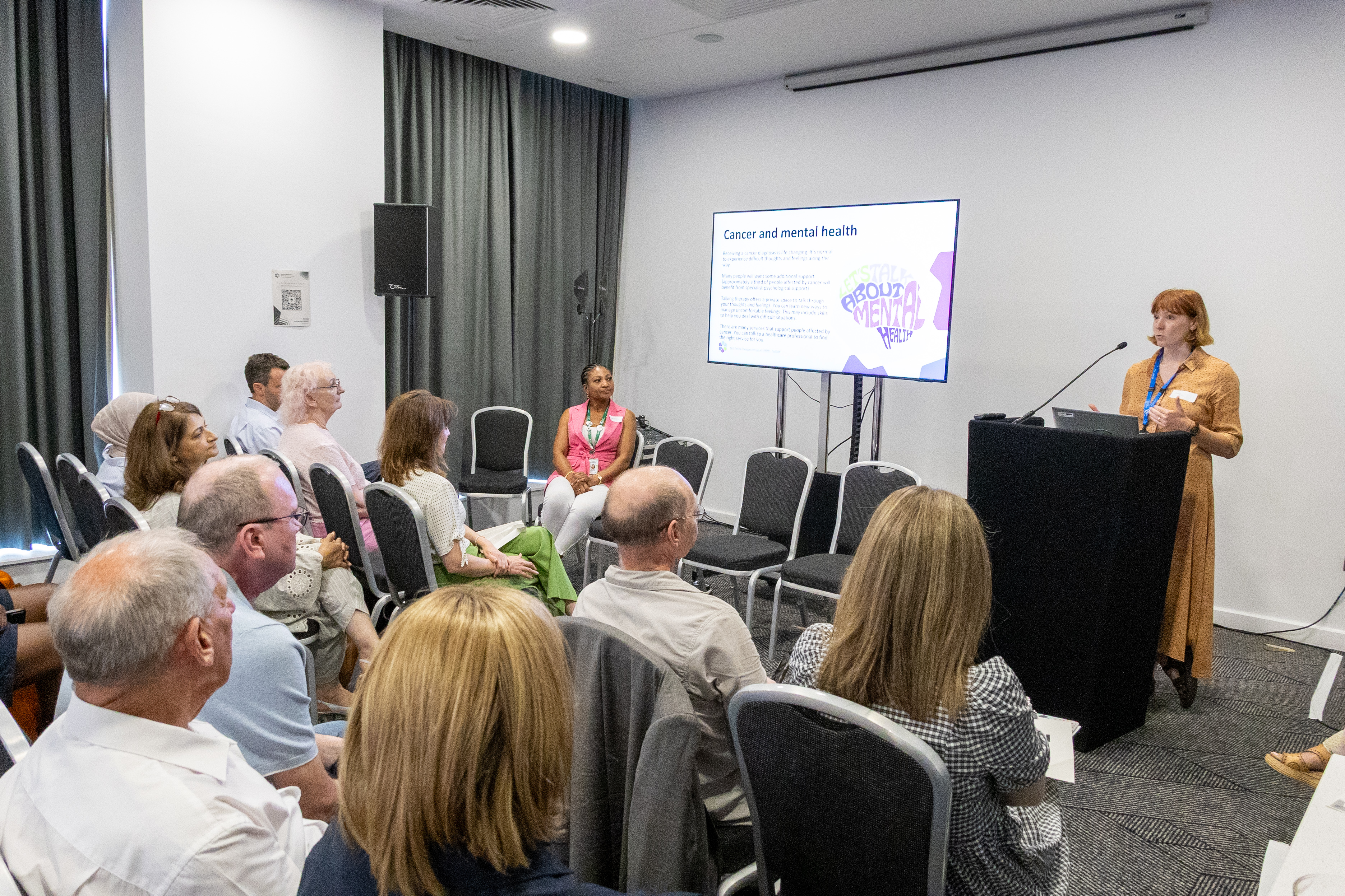 39 A group of people seated in rows is listening to a presentation in a conference room Sophie Bates stands at a podium beside a screen displaying a slide about "Cancer and mental health." The room has white walls, grey curtains, and a projector mounted on the ceiling.
