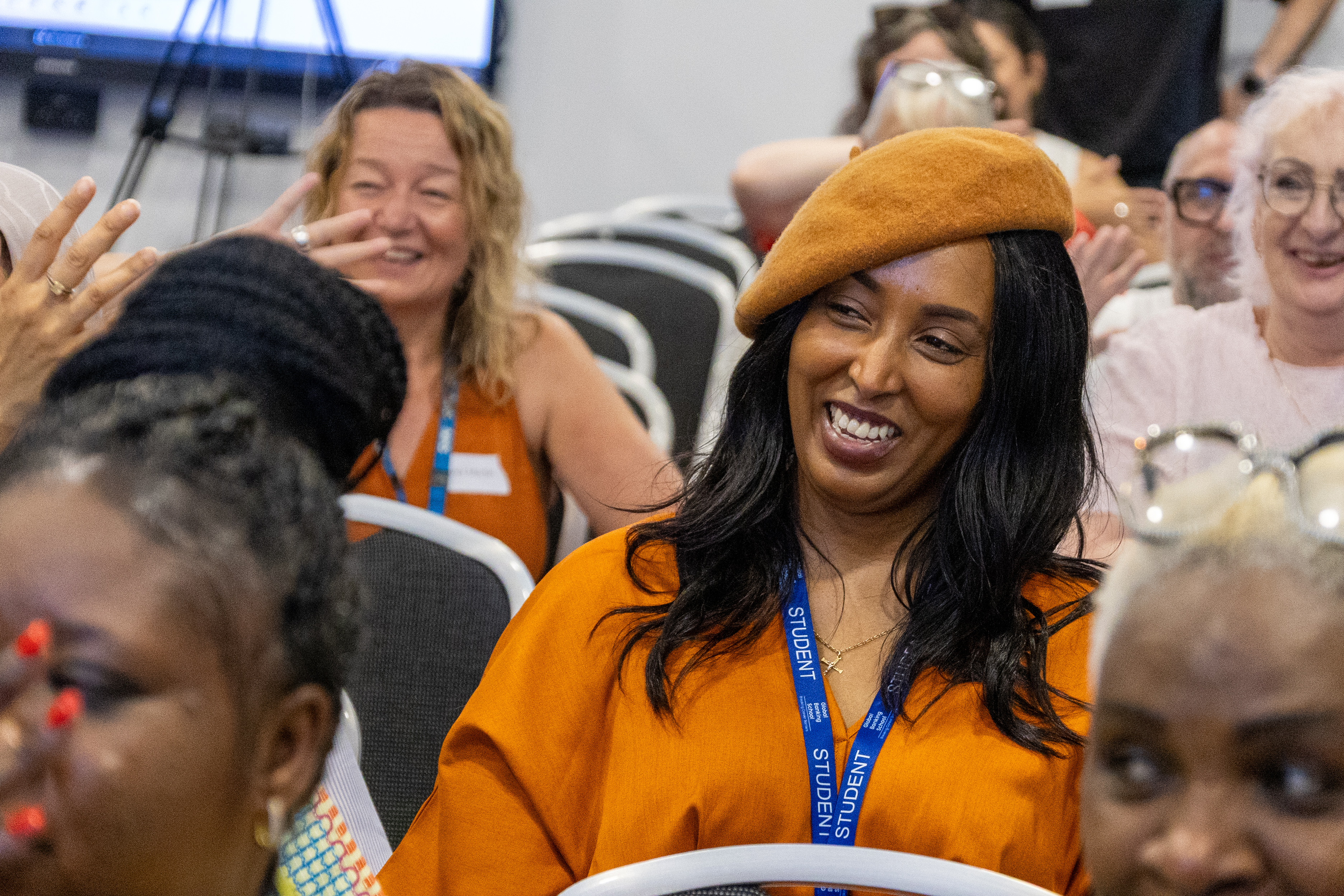 4 A group of people sitting in a conference room, smiling. One person in the foreground wears a bright orange outfit and a matching beret.