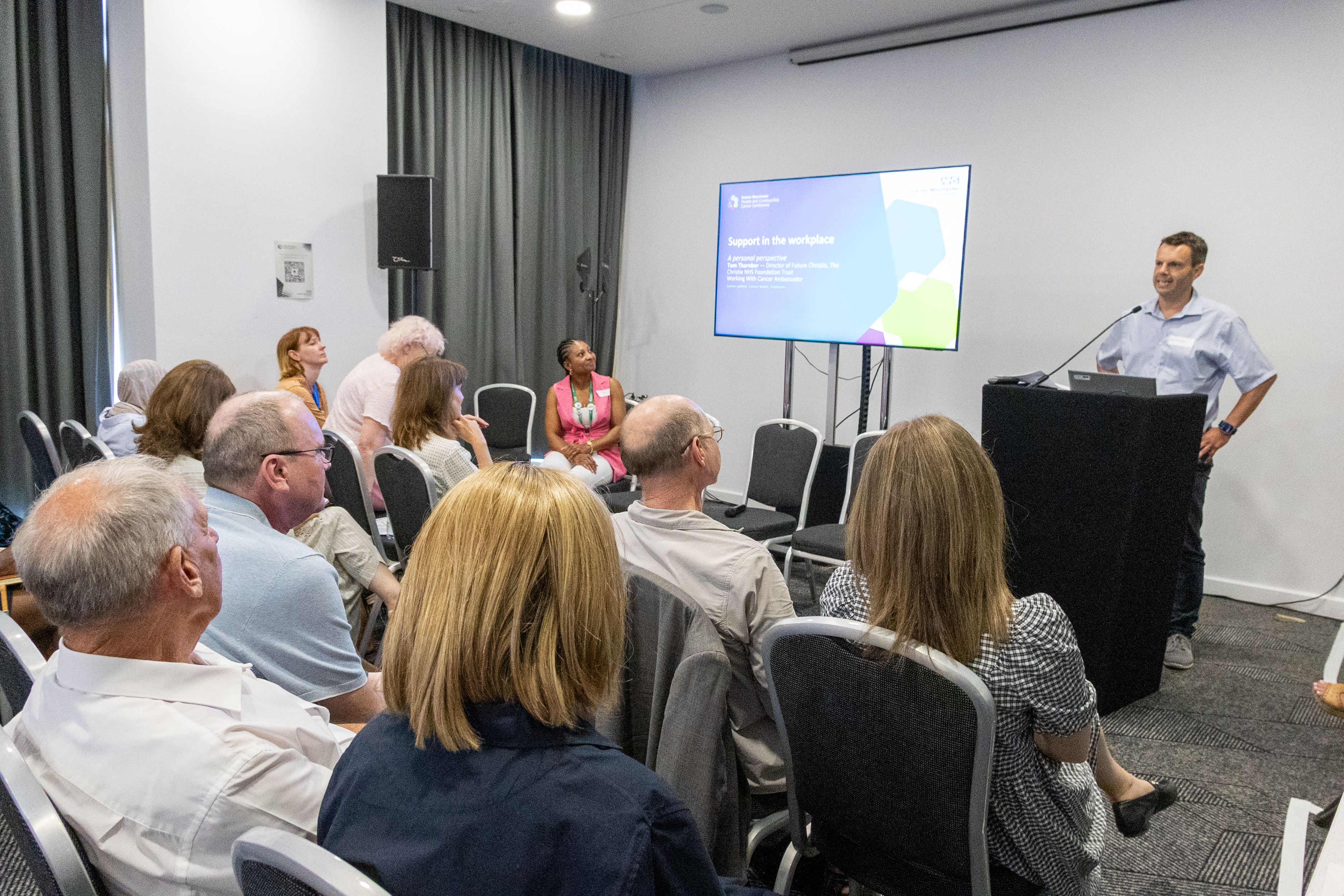 41 Tom Thornber stood presenting at the front of a conference behind a podium with hands on his hips addressing a room full of delegates about support in the workforce