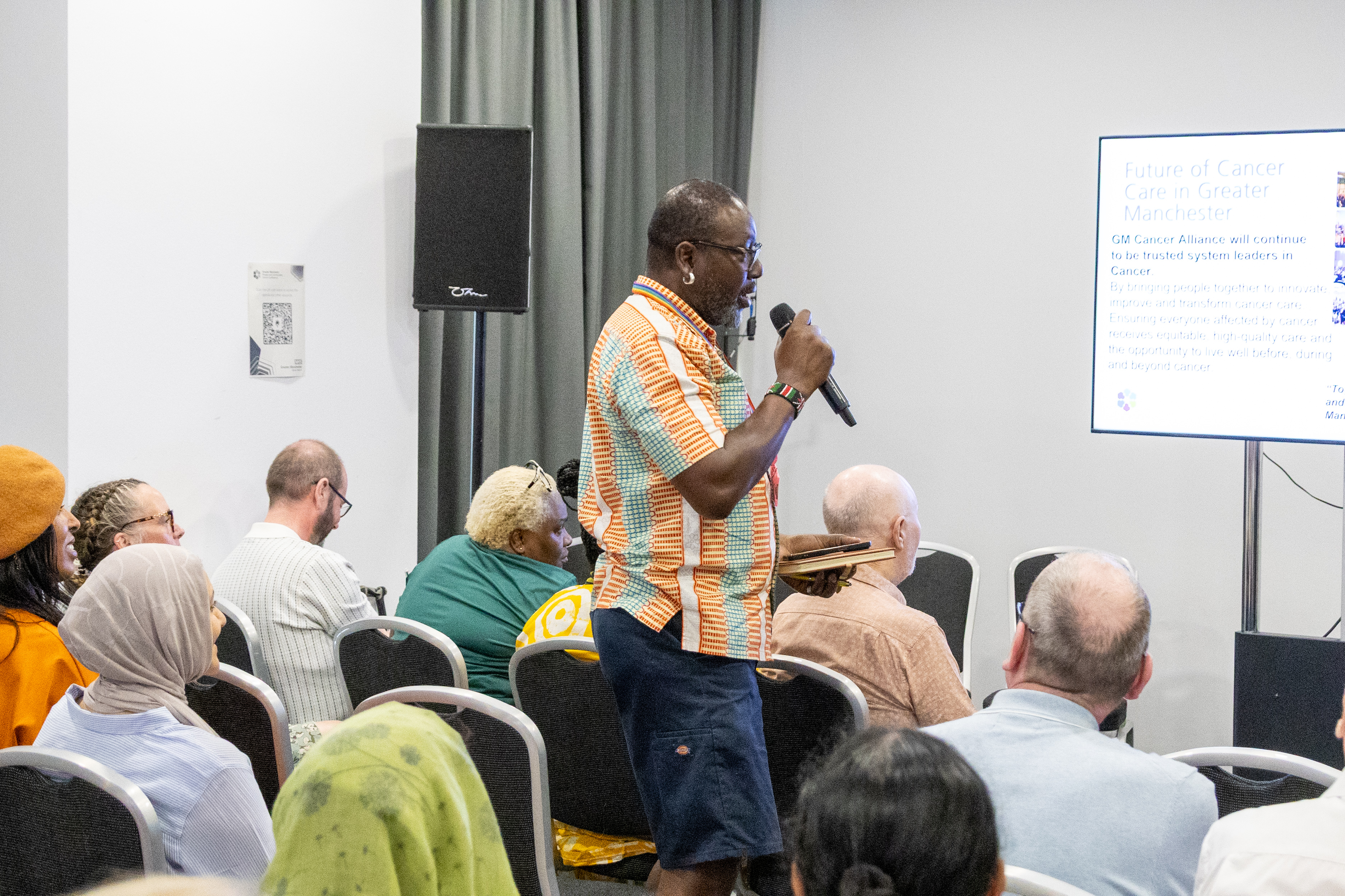 5 A person in a colourful shirt speaks into a microphone at a conference. The audience is seated, listening attentively. A screen displays a presentation titled "Future of Cancer Care in Greater Manchester.