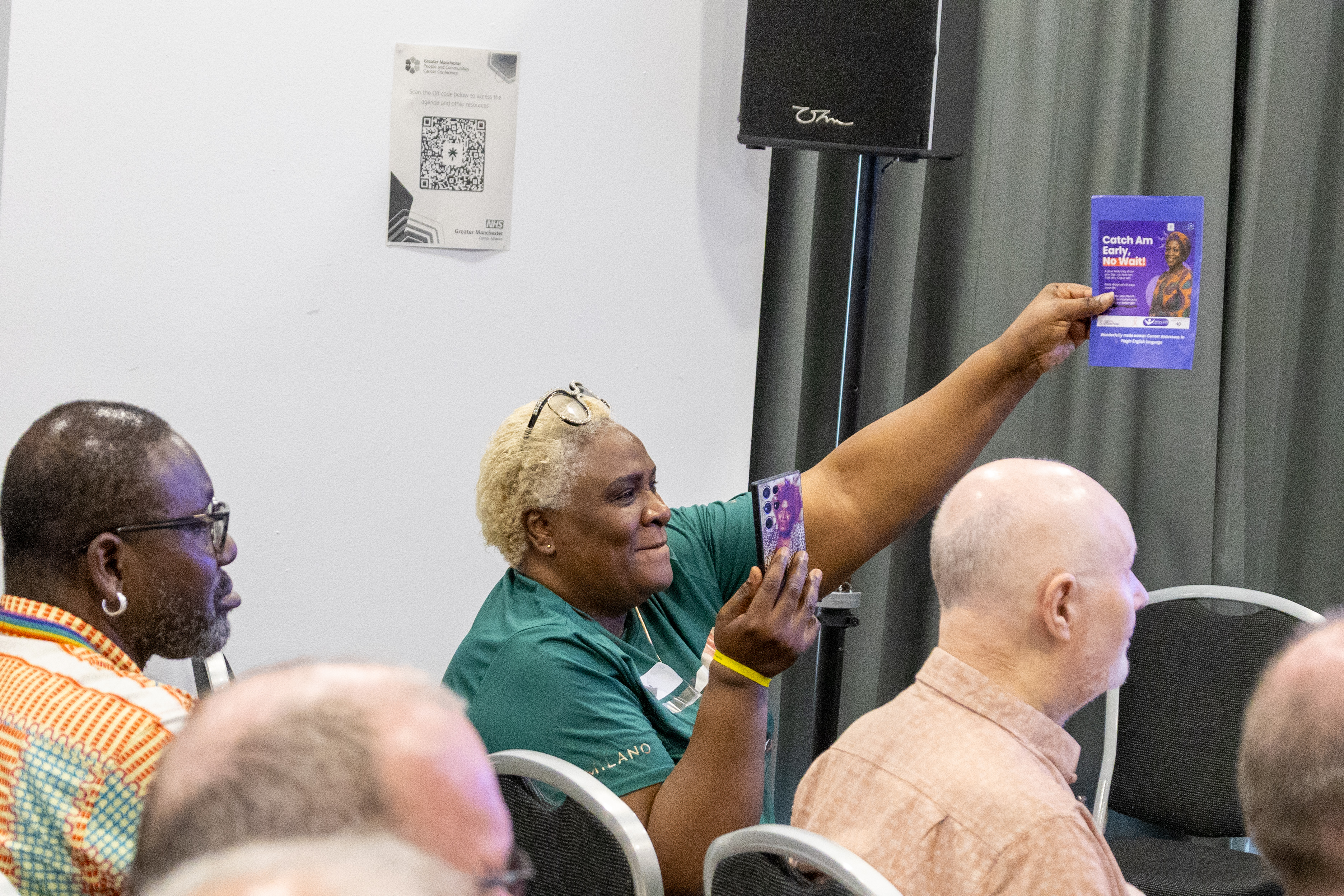 7 A person holds up a leaflet to show the delegates sitting in a conference setting. The room has a QR code poster on the wall.
