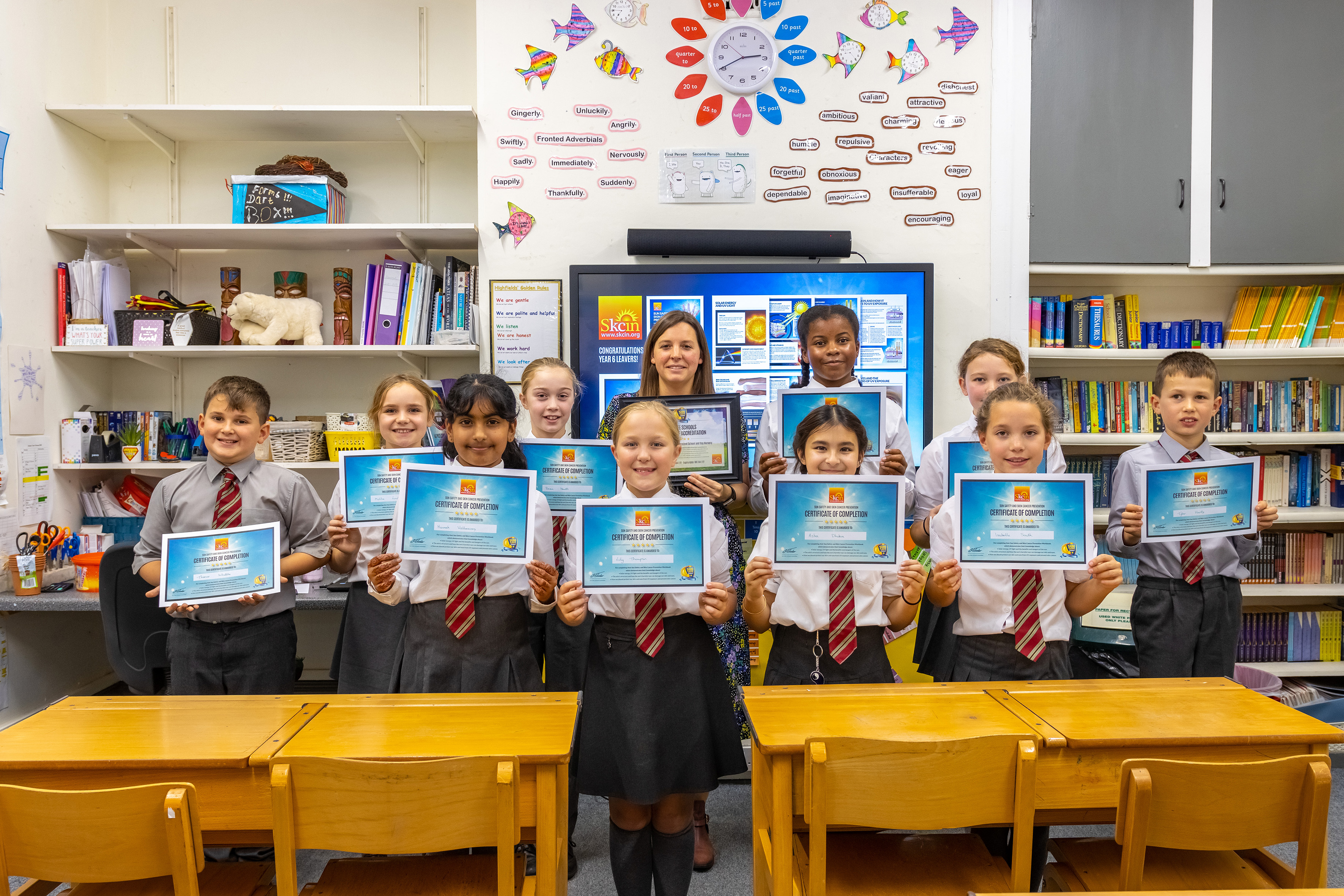 A school taking part in Sun Safe Schools 1 A group of primary school pupils stand in a classroom holding Sun Safe Schools certificates alongside their teacher after completing the Sun Safe Schools programme. A display about sun safety is visible on the screen behind them.