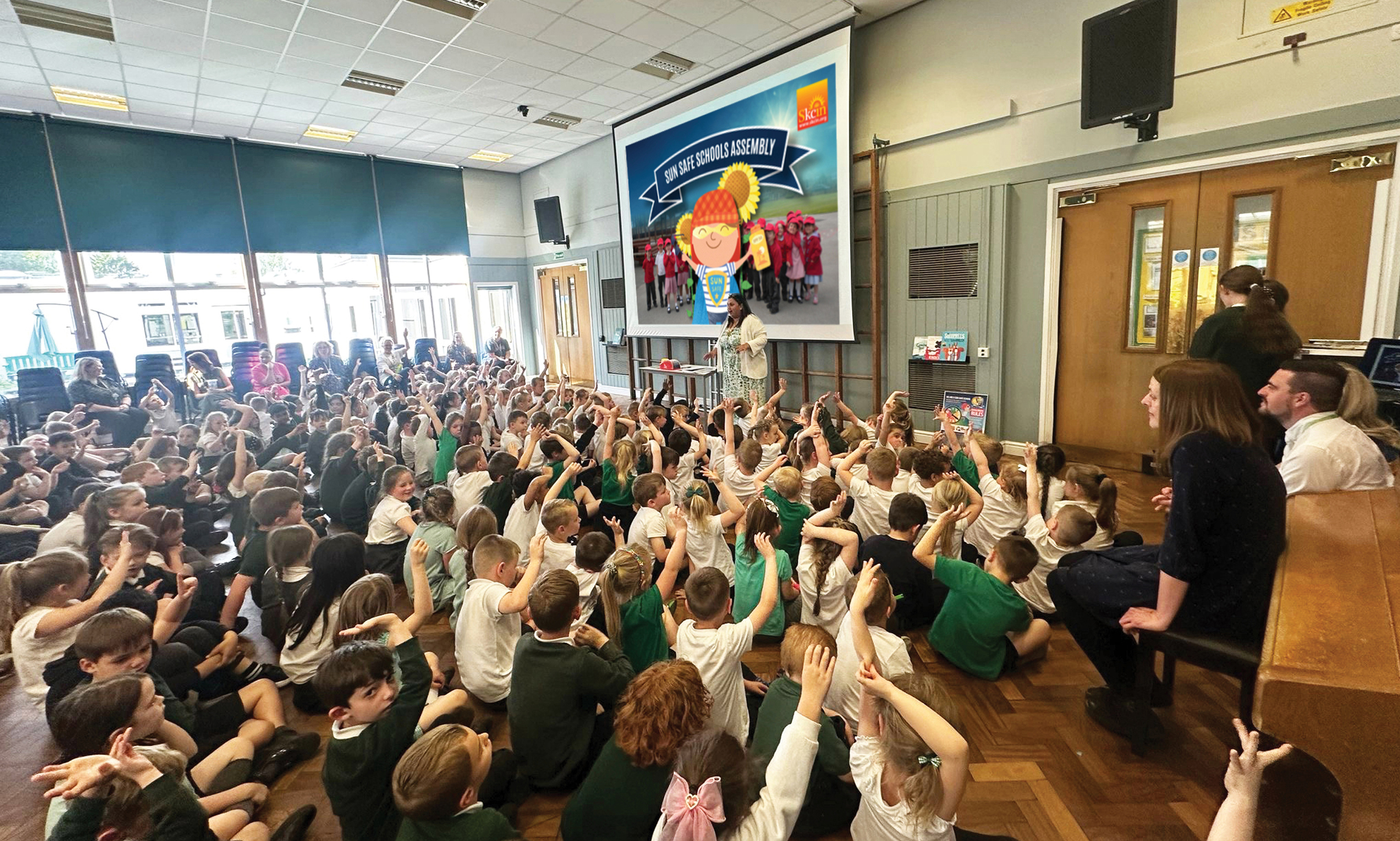 A school taking part in sun safe schools A school hall filled with primary school pupils sitting on the floor with their hands raised as they take part in an interactive Sun Safe Schools assembly. A presenter stands at the front beside a large screen displaying a sun safety presentation, while teachers watch from the side.