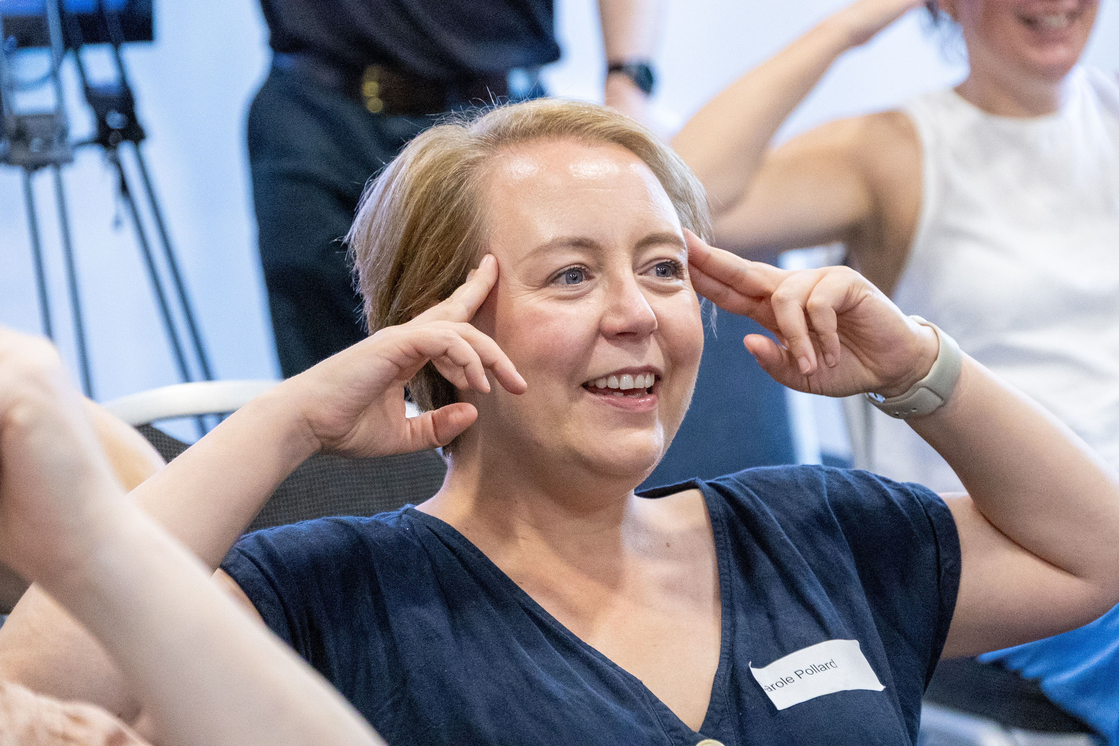 BR1A3986 Carole smiling with hands pointing to their temple at the People and Communities Conference joining in the chair yoga activity