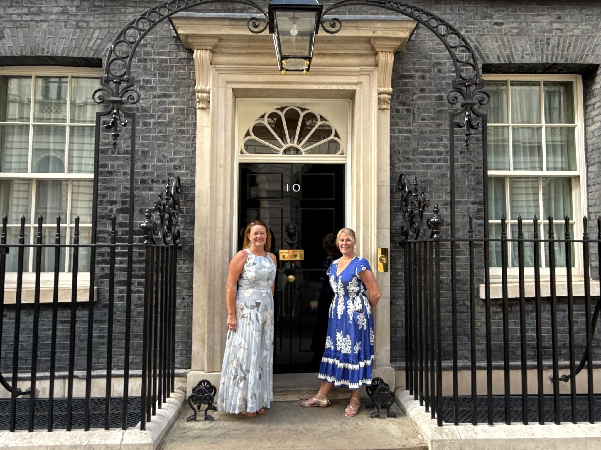 Downing Street Reception – Susi and Alison A Alison and Susannah wearing long dresses stand in front of the black door of Number 10 Downing Street