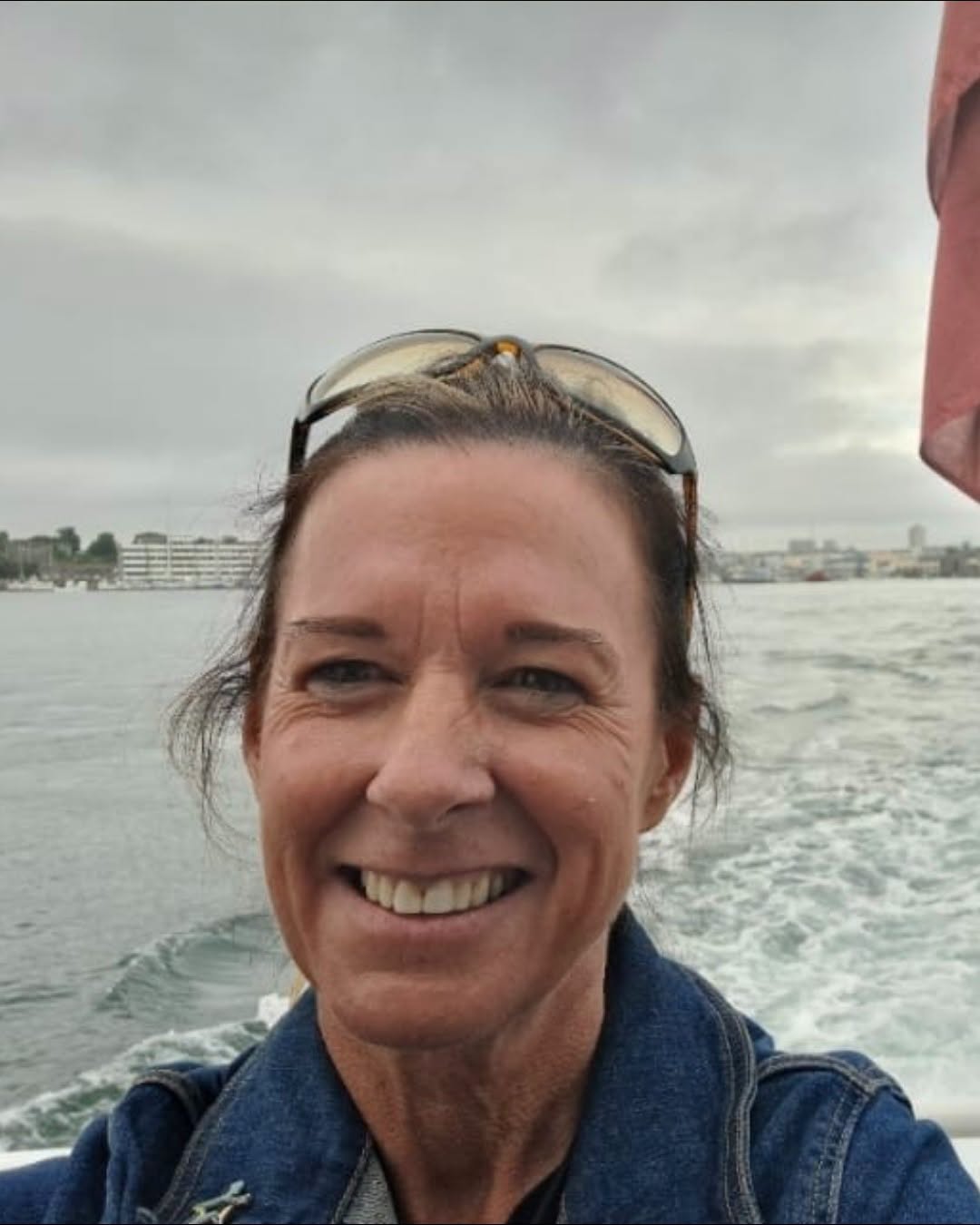 Joanne – Bowel cancer NHS E story Joanne a woman with brown hair tied back and sun glasses on her head and a beach in the background