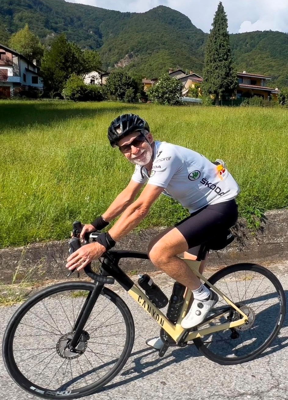 Pete - a white man in his 60s wearing a white T-shirt and cycle helmet cycles on a bike on a path close to grassy fields and with rolling hills in the background 
