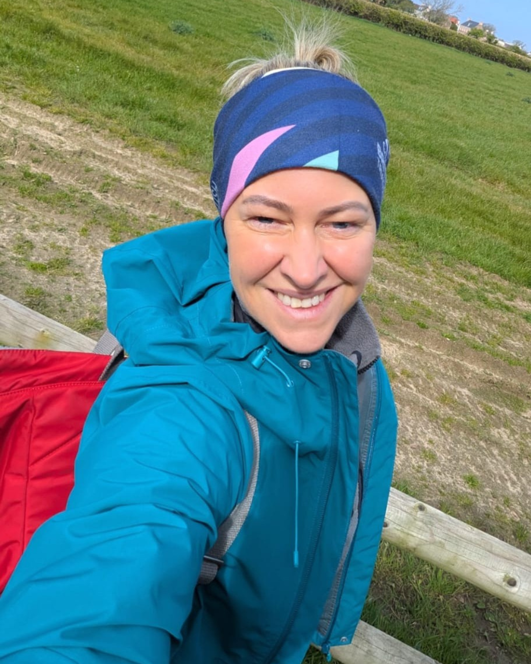 A person in a teal jacket and colourful headband takes a selfie outdoors with a red backpack visible. They are standing near a wooden fence with a green field and houses in the background under a clear blue sky.